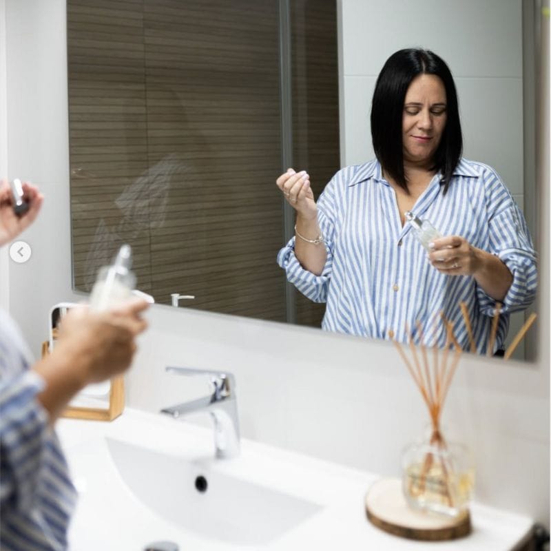 Paqui en el baño, reflejada en el espejo perfumando con un spray de ambiente y un mikado.